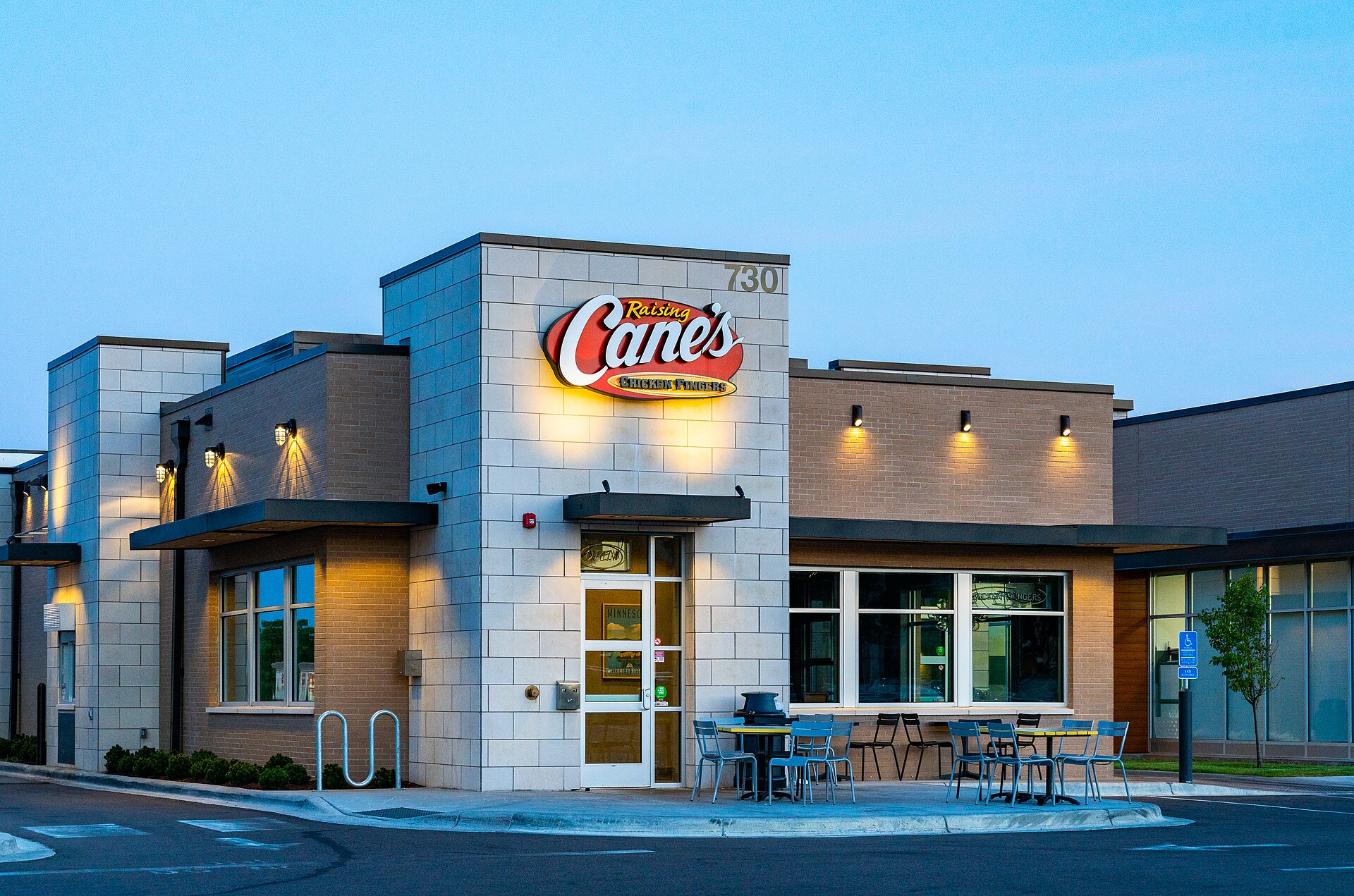 Photo of a typical Raising Cane's Chicken Fingers restaurant at dusk with its lit sign and patio seating, in Woodbury, Minnesota
